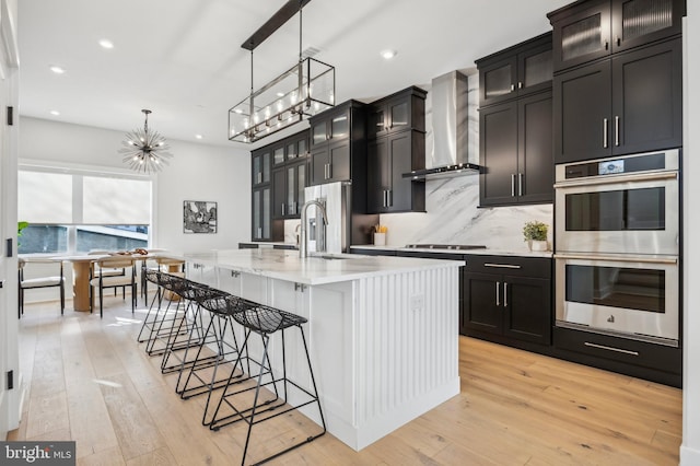 kitchen featuring a center island with sink, stainless steel appliances, backsplash, hanging light fixtures, and wall chimney exhaust hood