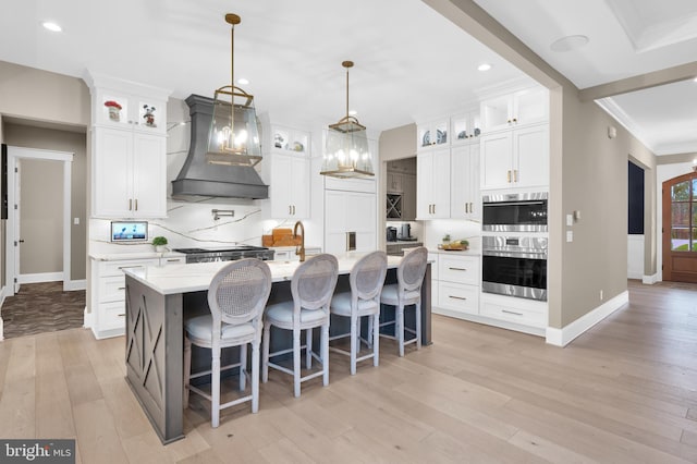 kitchen featuring white cabinets, an island with sink, custom range hood, and pendant lighting