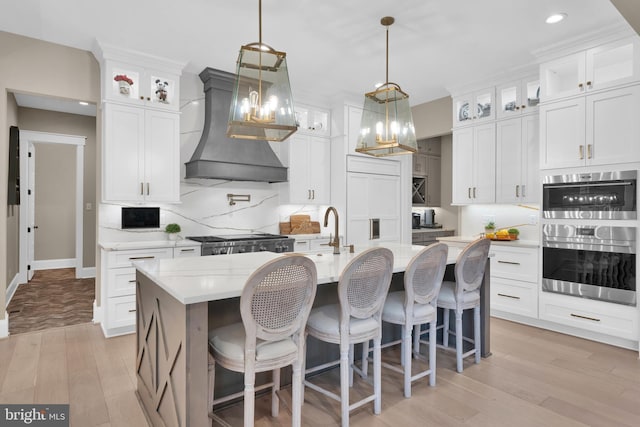 kitchen with premium range hood, white cabinetry, stove, decorative light fixtures, and a center island with sink