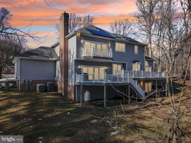 back house at dusk with a lawn, a wooden deck, solar panels, central air condition unit, and a balcony