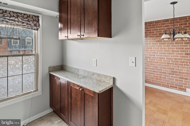 kitchen with dark brown cabinets, hanging light fixtures, light stone counters, brick wall, and a notable chandelier