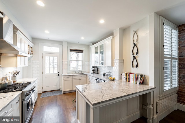 kitchen featuring wall chimney exhaust hood, light stone counters, kitchen peninsula, stainless steel appliances, and white cabinets