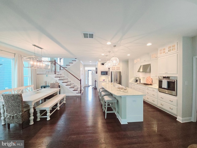 kitchen featuring pendant lighting, wall chimney range hood, appliances with stainless steel finishes, and a kitchen island with sink