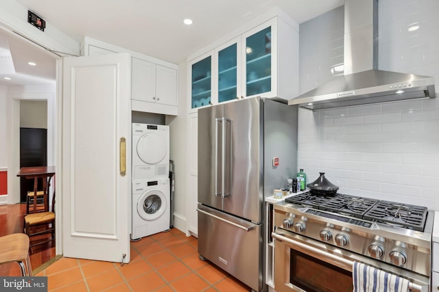 kitchen with appliances with stainless steel finishes, wall chimney exhaust hood, backsplash, white cabinets, and stacked washer and clothes dryer