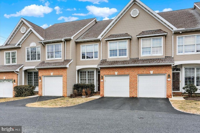 townhome / multi-family property with driveway, a shingled roof, a garage, and brick siding