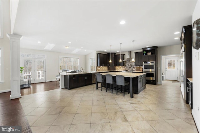 kitchen with wall chimney exhaust hood, ornate columns, an island with sink, pendant lighting, and stainless steel appliances