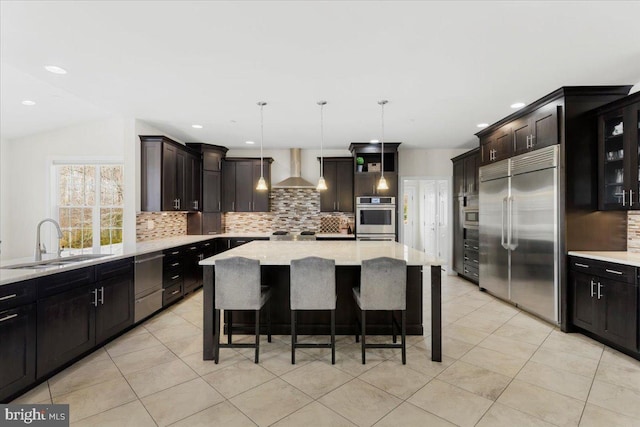 kitchen with sink, hanging light fixtures, stainless steel appliances, a kitchen island, and wall chimney exhaust hood