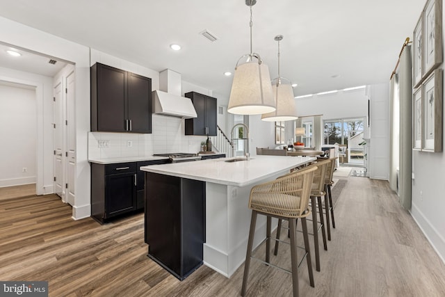 kitchen with hardwood / wood-style floors, pendant lighting, wall chimney exhaust hood, decorative backsplash, and a kitchen island with sink