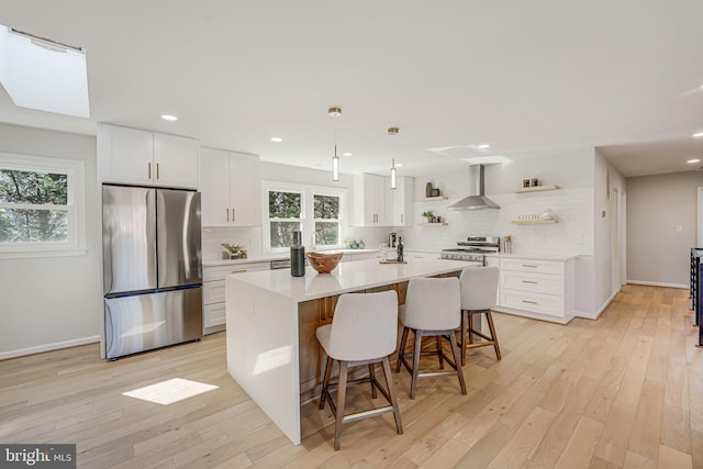 kitchen with decorative light fixtures, white cabinets, stainless steel appliances, a center island with sink, and wall chimney exhaust hood