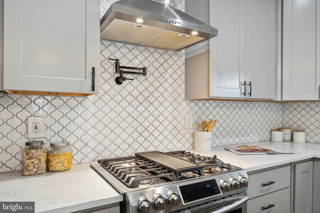 kitchen featuring wall chimney exhaust hood, gray cabinetry, gas range, light stone counters, and backsplash