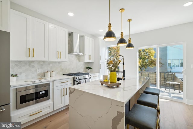 kitchen featuring light stone countertops, white cabinets, appliances with stainless steel finishes, wall chimney range hood, and a center island with sink