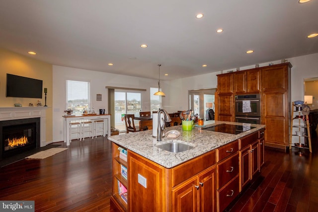 kitchen featuring sink, hanging light fixtures, light stone counters, black electric stovetop, and a center island with sink