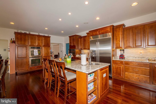 kitchen featuring appliances with stainless steel finishes, a breakfast bar, light stone counters, dark wood-type flooring, and a center island with sink