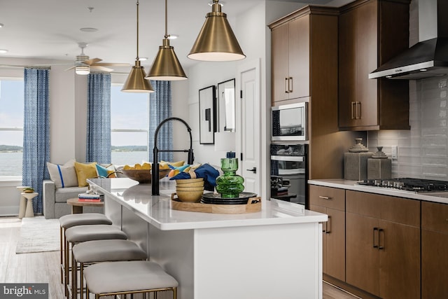 kitchen featuring wall chimney exhaust hood, stainless steel appliances, backsplash, a kitchen island with sink, and a breakfast bar