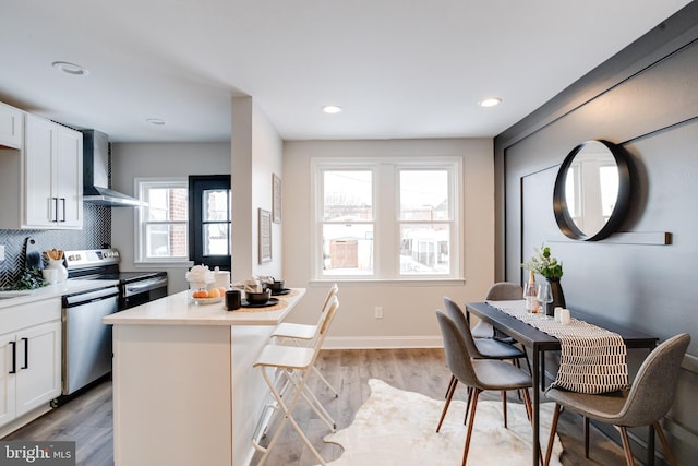 kitchen featuring wall chimney range hood, light wood-type flooring, white cabinets, and appliances with stainless steel finishes