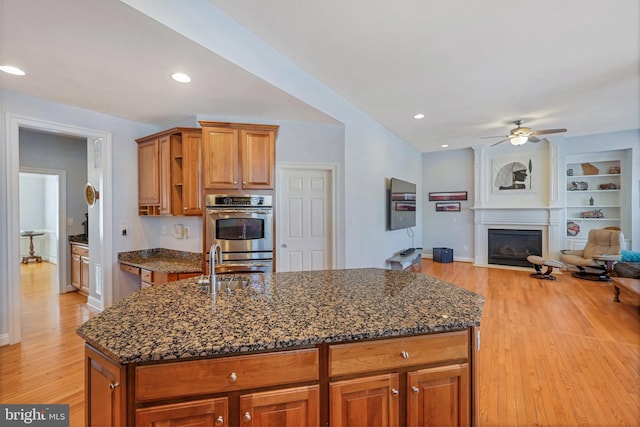 kitchen with dark stone countertops, stainless steel double oven, a center island with sink, light wood-type flooring, and built in shelves