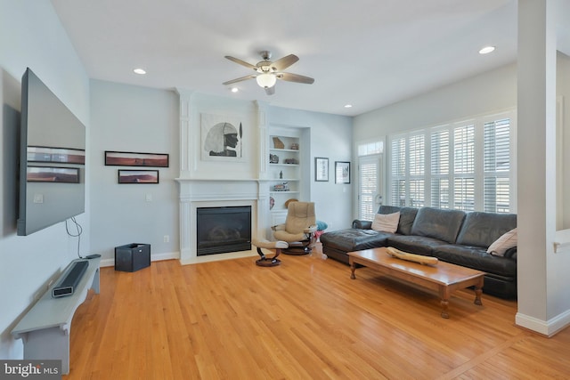 living room with built in shelves, a large fireplace, ceiling fan, and light wood-type flooring