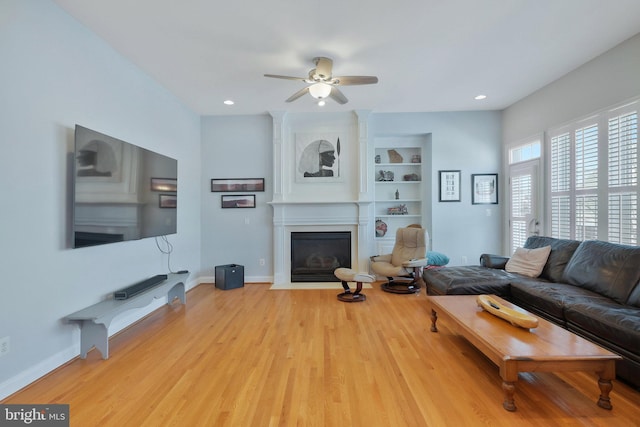 living room with built in shelves, a large fireplace, ceiling fan, and light hardwood / wood-style flooring