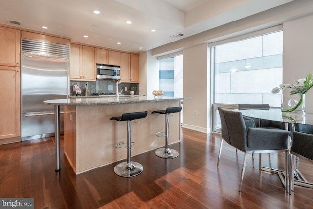 kitchen featuring stone countertops, light brown cabinetry, backsplash, a kitchen island with sink, and stainless steel appliances