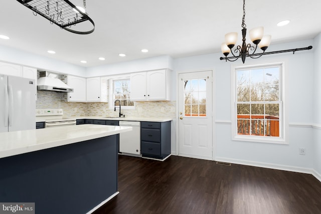 kitchen featuring wall chimney range hood, white appliances, sink, white cabinetry, and hanging light fixtures