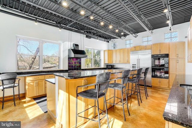 kitchen with a towering ceiling, finished concrete floors, under cabinet range hood, light brown cabinets, and built in fridge