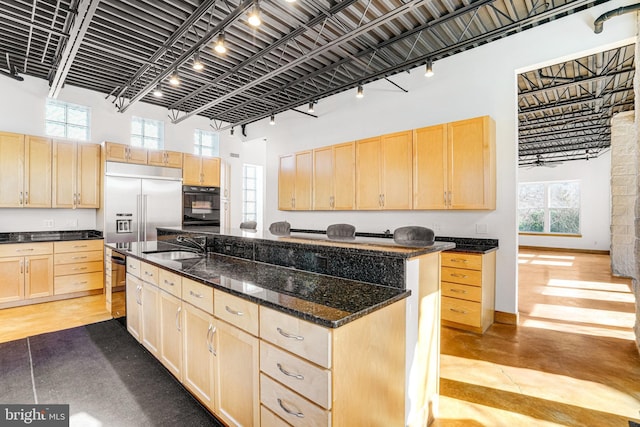 kitchen with a towering ceiling, an island with sink, oven, built in refrigerator, and light brown cabinetry