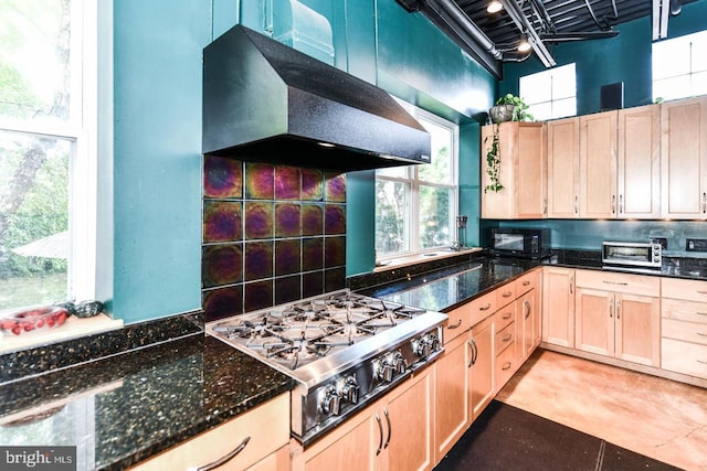 kitchen with dark stone counters, stainless steel gas stovetop, under cabinet range hood, light brown cabinets, and backsplash