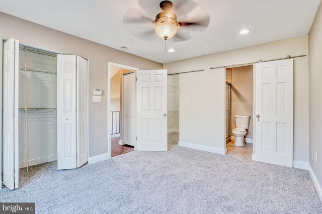 unfurnished bedroom featuring two closets, carpet flooring, ceiling fan, ensuite bathroom, and a barn door