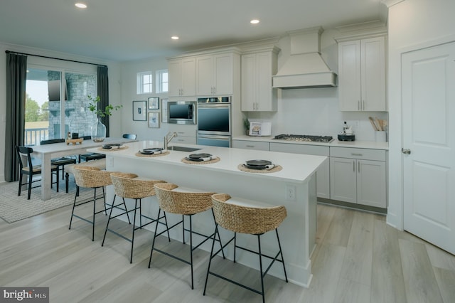 kitchen with appliances with stainless steel finishes, a breakfast bar, an island with sink, white cabinetry, and custom exhaust hood