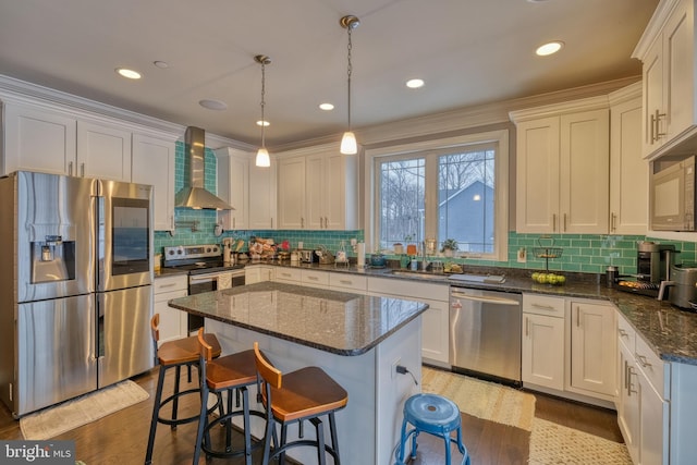 kitchen featuring wall chimney range hood, stainless steel appliances, a kitchen island, decorative light fixtures, and dark stone counters