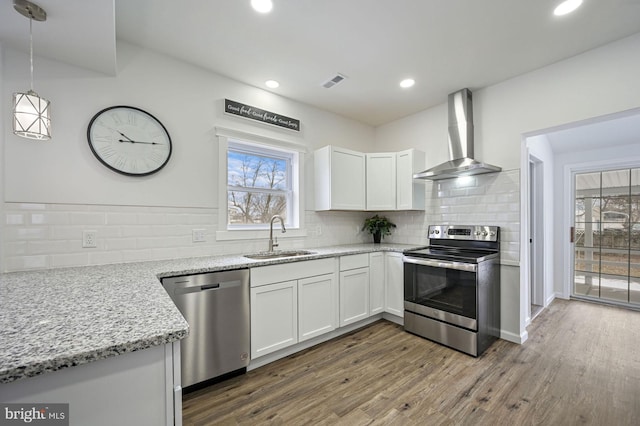 kitchen featuring white cabinets, wall chimney range hood, stainless steel appliances, sink, and hanging light fixtures