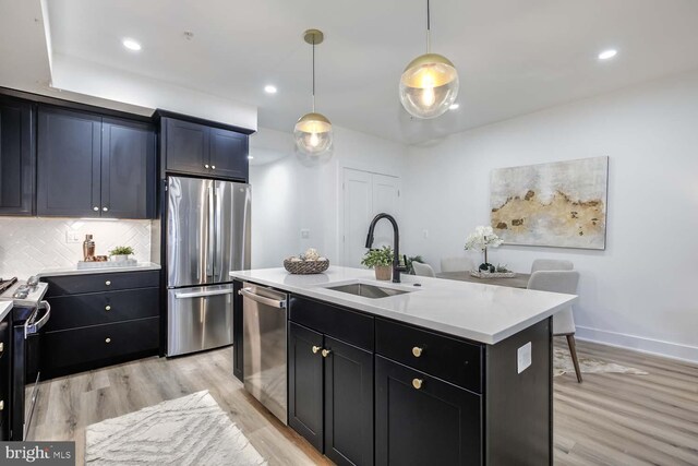 kitchen featuring appliances with stainless steel finishes, sink, light wood-type flooring, decorative light fixtures, and an island with sink