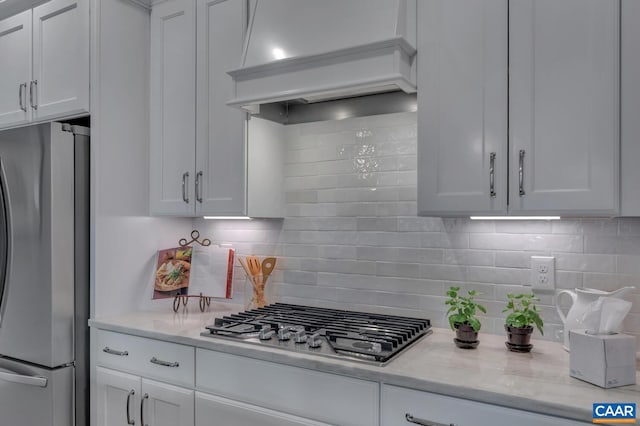 kitchen featuring light stone countertops, appliances with stainless steel finishes, premium range hood, decorative backsplash, and white cabinets