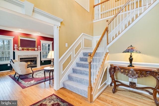 staircase featuring crown molding, hardwood / wood-style floors, wainscoting, a high ceiling, and a glass covered fireplace