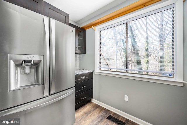 kitchen with stainless steel refrigerator with ice dispenser, dark brown cabinetry, wood-type flooring, and light stone counters