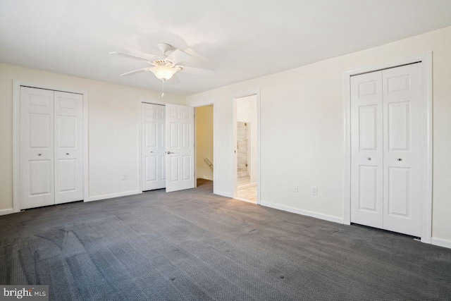 unfurnished bedroom with ceiling fan, two closets, dark carpet, and ensuite bath