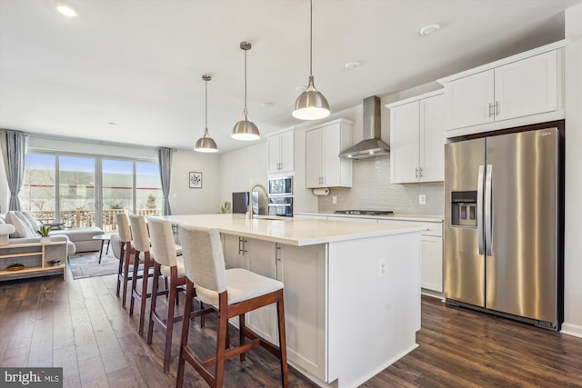 kitchen with hanging light fixtures, a center island with sink, appliances with stainless steel finishes, wall chimney range hood, and white cabinets