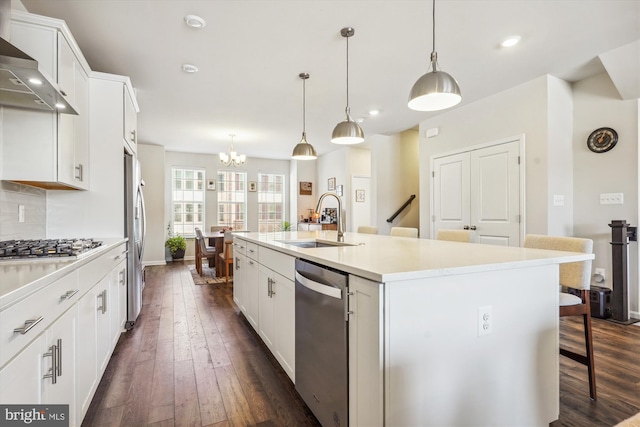 kitchen with an island with sink, sink, hanging light fixtures, stainless steel appliances, and wall chimney range hood