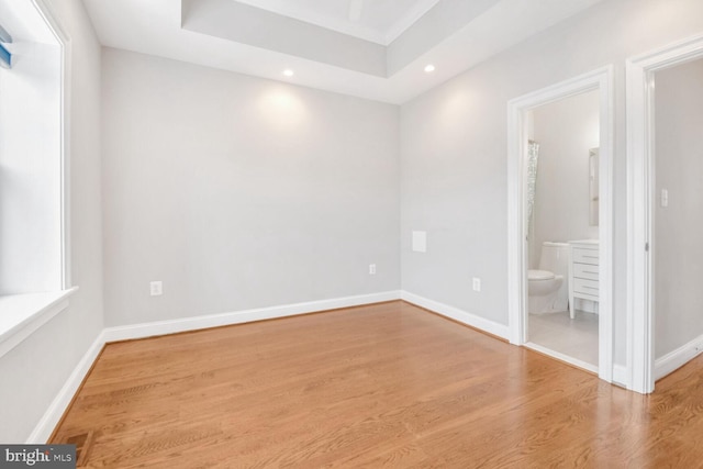 unfurnished bedroom featuring light hardwood / wood-style floors, multiple windows, ensuite bath, and a raised ceiling