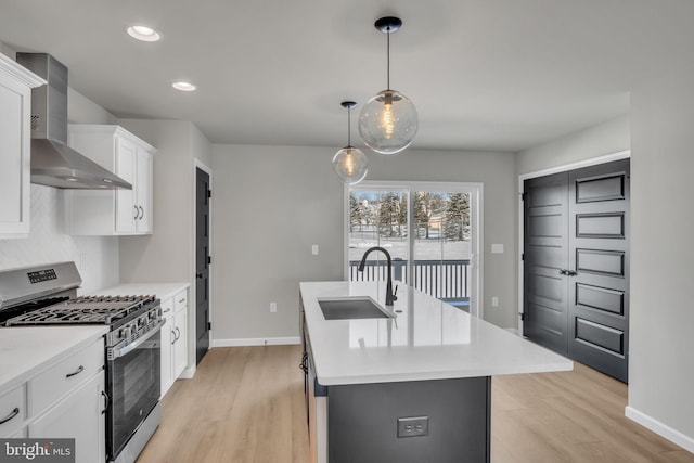 kitchen with wall chimney range hood, sink, white cabinetry, stainless steel gas range oven, and a center island with sink