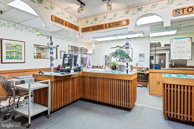 kitchen with carpet flooring, kitchen peninsula, a drop ceiling, and wood walls