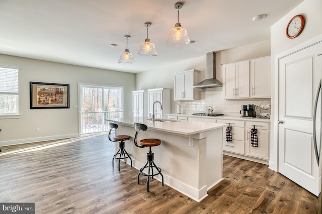 kitchen with wall chimney exhaust hood, sink, a center island with sink, hanging light fixtures, and white cabinets