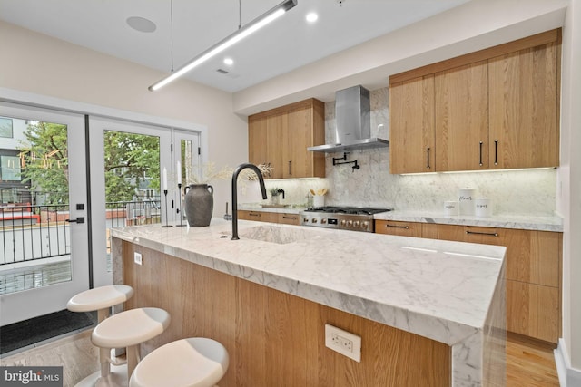 kitchen with wall chimney range hood, an island with sink, tasteful backsplash, sink, and light stone counters