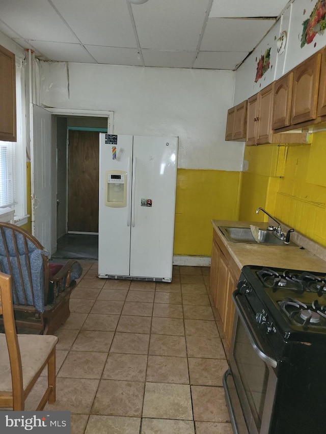 kitchen with sink, stainless steel gas stove, light tile patterned floors, white refrigerator with ice dispenser, and a drop ceiling