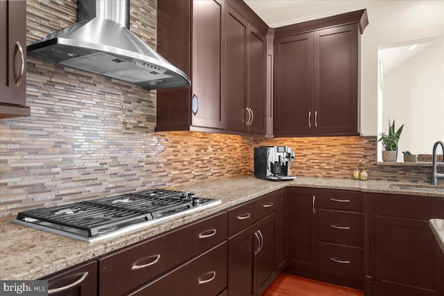 kitchen featuring sink, backsplash, stainless steel gas cooktop, light stone counters, and wall chimney range hood