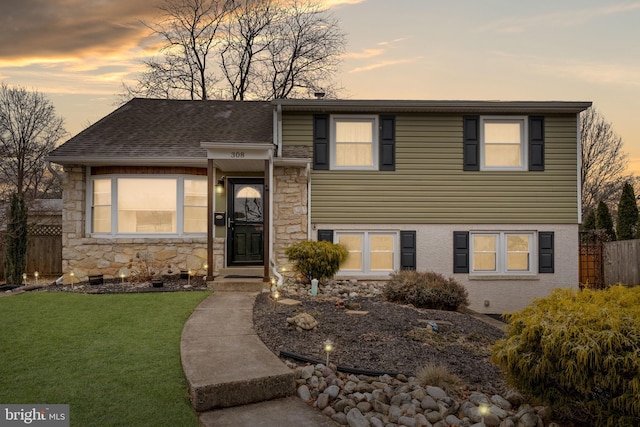 tri-level home with stone siding, fence, a front lawn, and roof with shingles