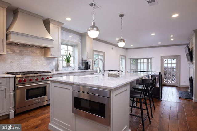 kitchen with hanging light fixtures, custom exhaust hood, light stone counters, a center island with sink, and stainless steel appliances