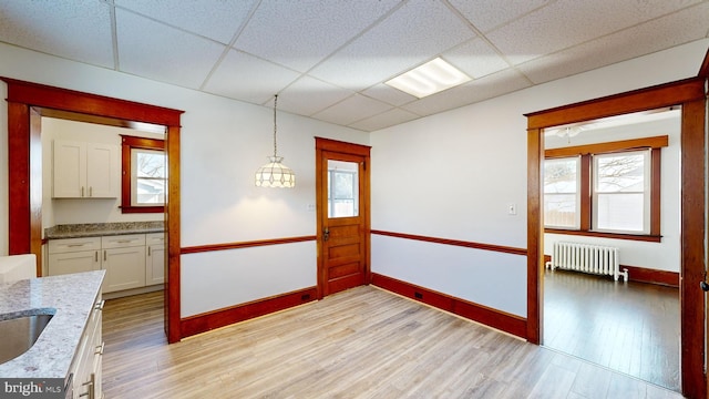 interior space with a paneled ceiling, radiator heating unit, white cabinets, hanging light fixtures, and light wood-type flooring