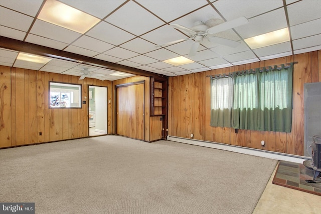 below grade area with a ceiling fan, carpet floors, a wood stove, wood walls, and baseboard heating