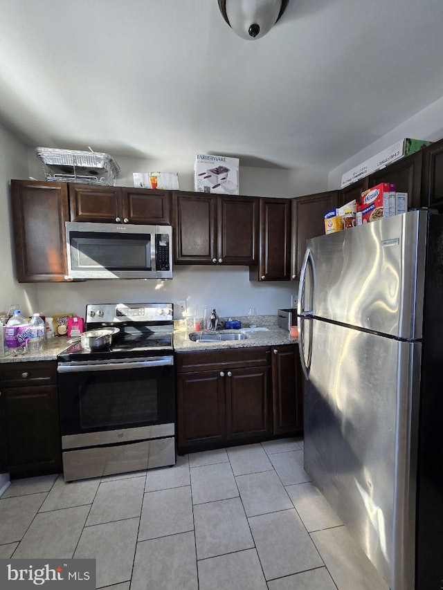 kitchen featuring dark brown cabinetry, sink, light tile patterned floors, appliances with stainless steel finishes, and light stone countertops